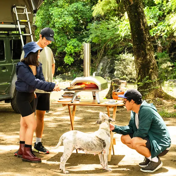 Drie mensen en een hond bij een Ooni pizzaoven buiten Drie mensen en een hond bij een Ooni pizzaoven buiten