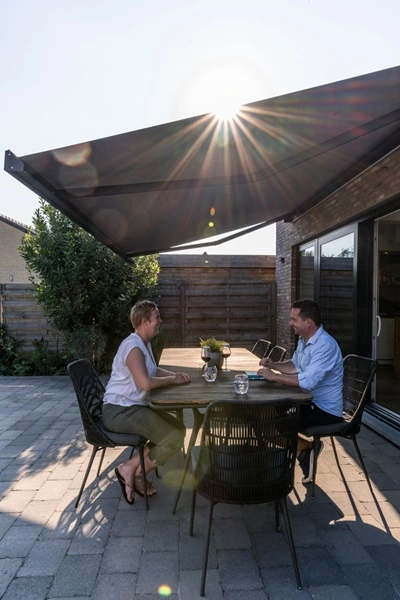BX270 knikarmscherm boven terras met twee mensen aan tafel, zonnestralen door doek heen BX270 knikarmscherm boven terras met twee mensen aan tafel, zonnestralen door doek heen