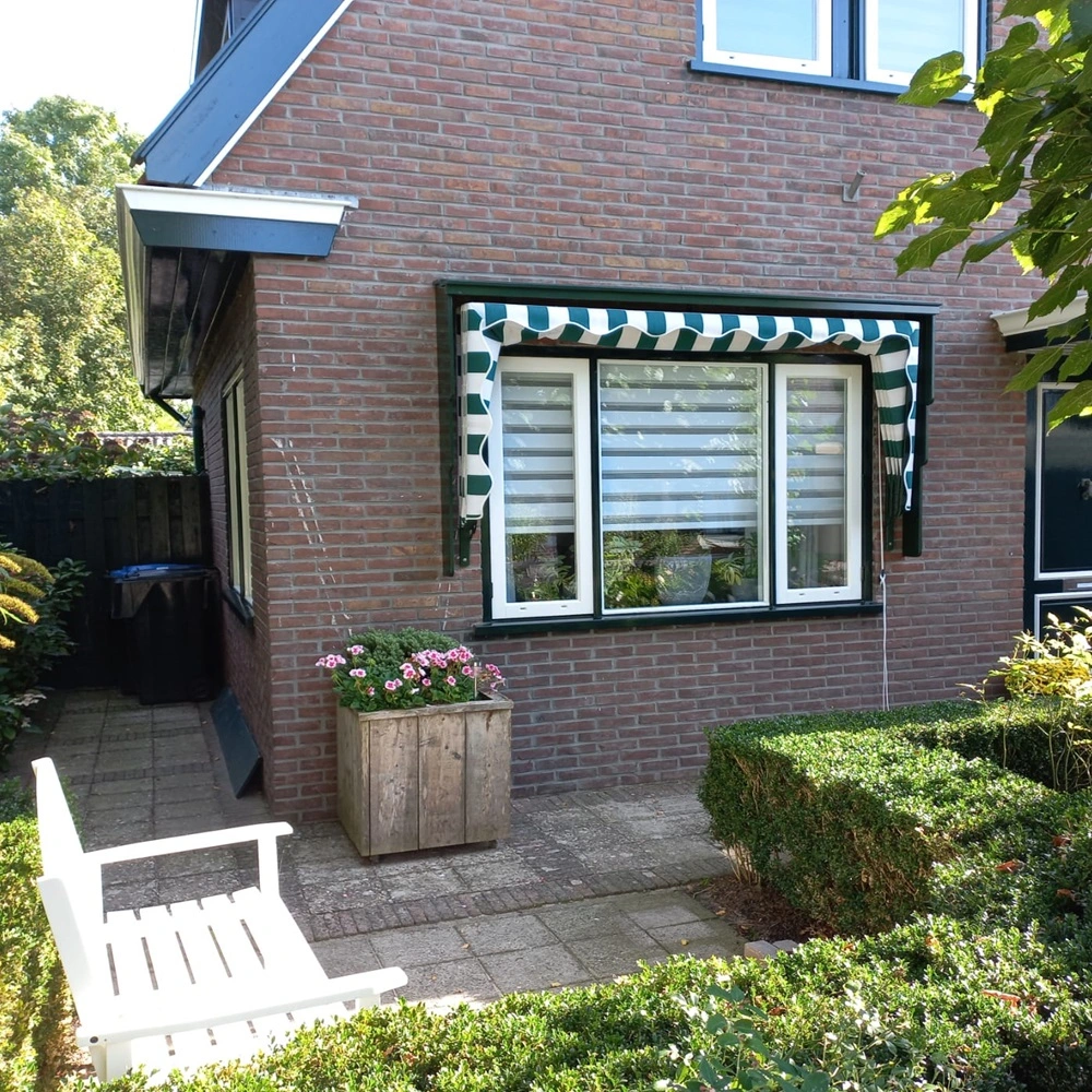 Aluminium markies met groene en witte strepen boven een raam naast een tuinpad