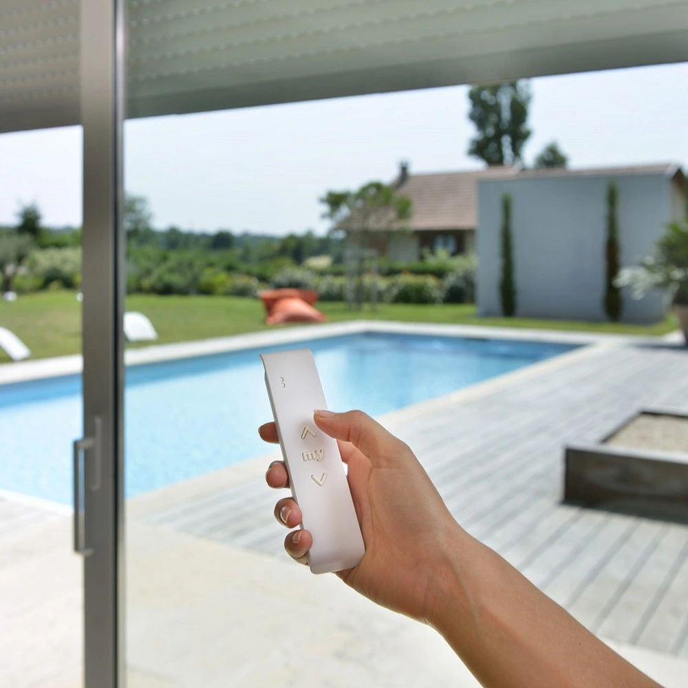Hand met afstandsbediening bedient zonwering met uitzicht op tuin en zwembad