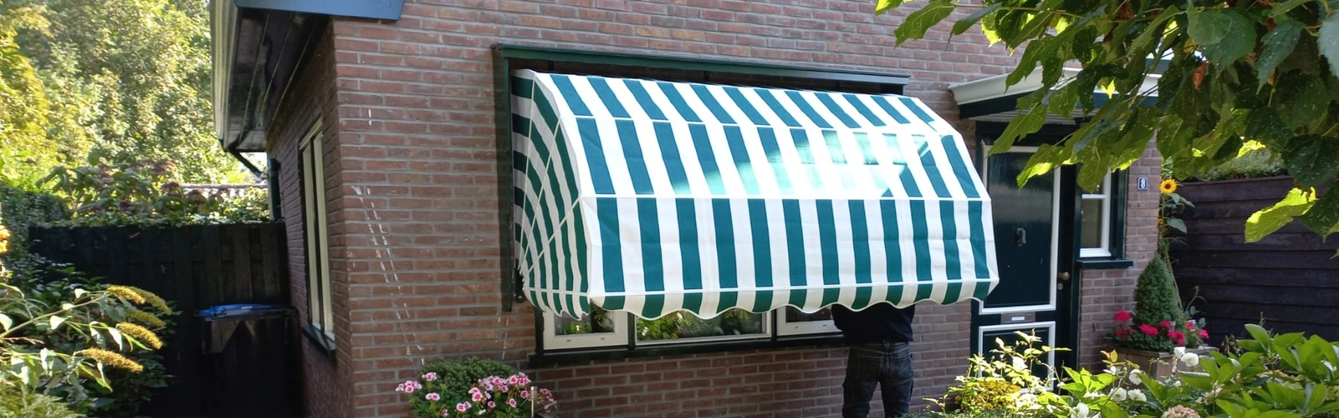 Groene en witte markies geopend boven een raam in een verzorgde tuin