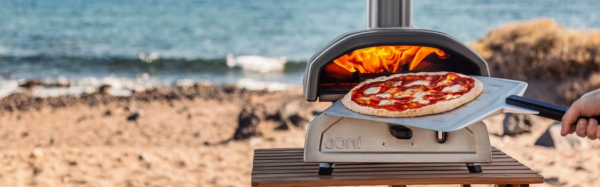 Houtgestookte Ooni pizza oven met pizza op houten tafel bij strand